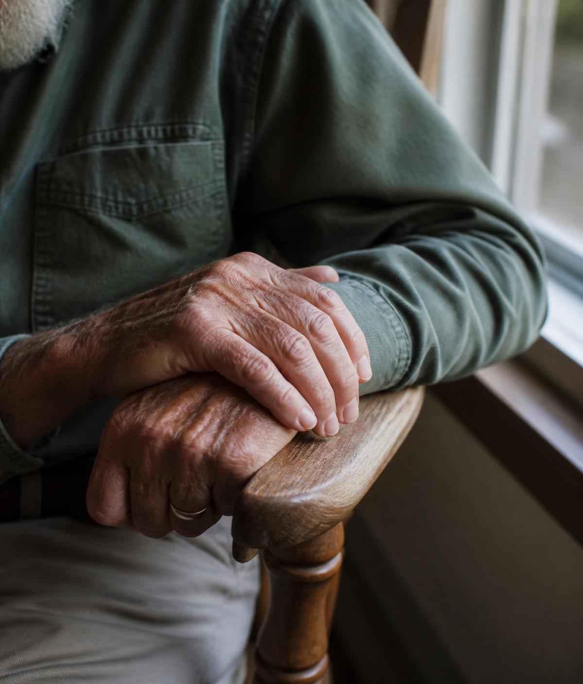 The hands of an elderly man resting gently on a wooden chair